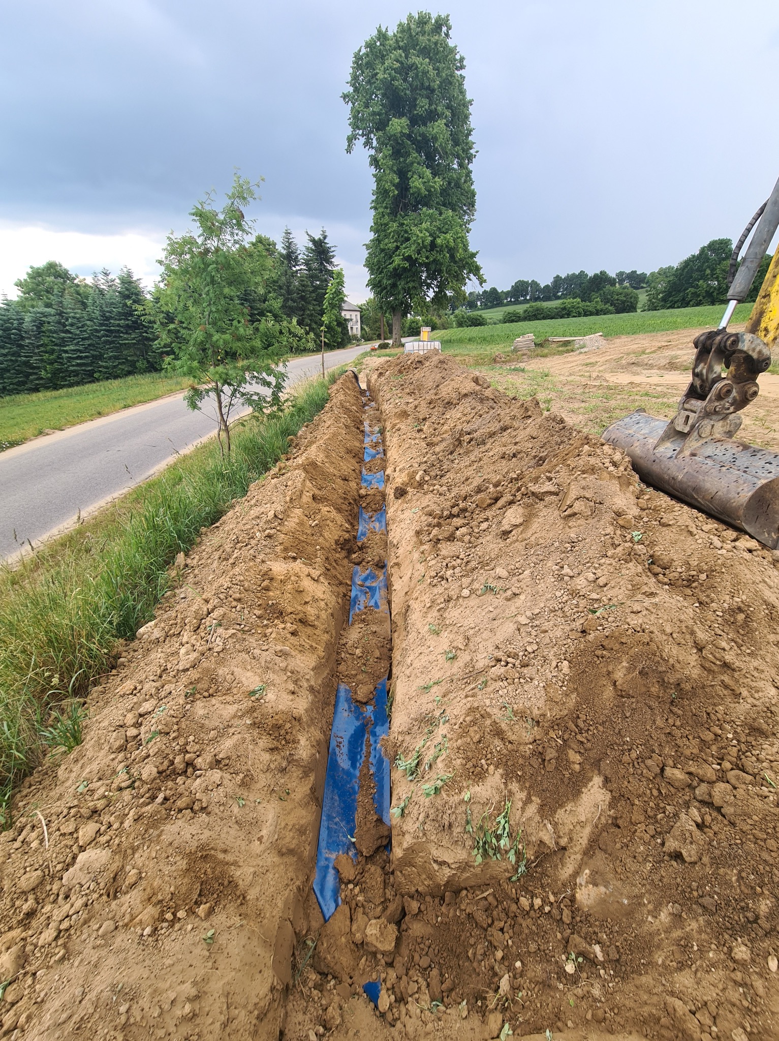 Wykopany rów w ziemi z niebieską folią ochronną, widoczna krawędź drogi asfaltowej i fragment koparki na tle zielonych drzew i pochmurnego nieba.