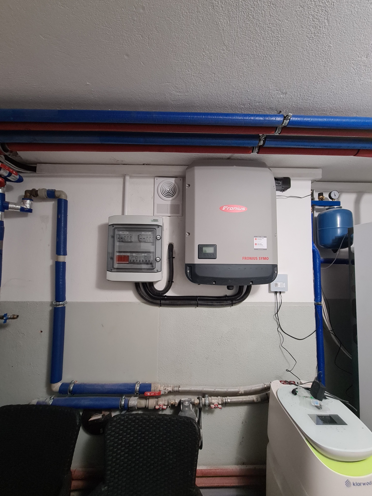 Szary inwerter Fronius Symo zamontowany na ścianie w pomieszczeniu technicznym, obok skrzynka z bezpiecznikami i niebieskie rury instalacji wodnej.