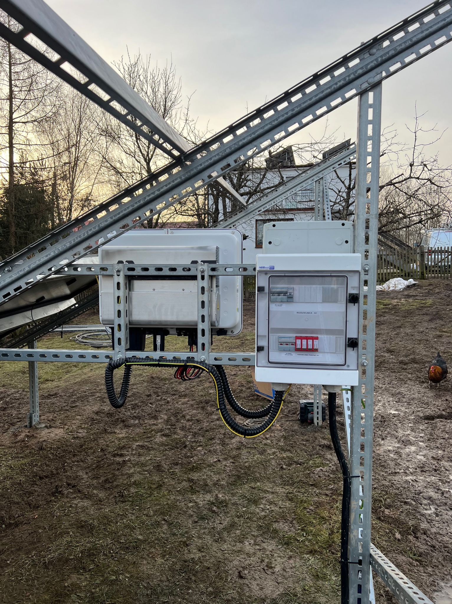 Instalacja fotowoltaiczna w Kartuzach w trakcie montażu: widoczna konstrukcja wsporcza, inwerter oraz rozdzielnica AC na tle błotnistego trawnika i zabudowań.