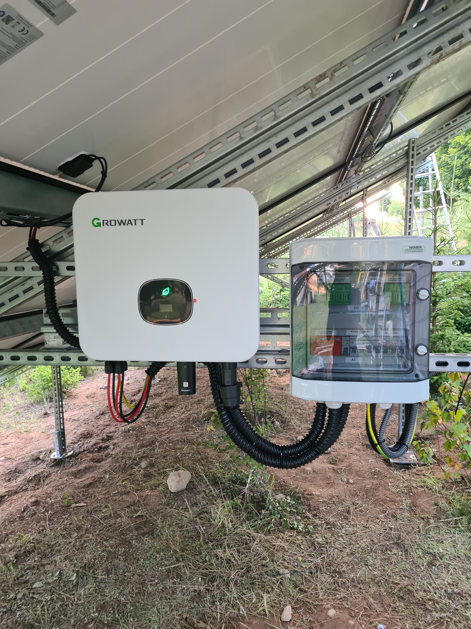 Instalacja fotowoltaiczna: biały inwerter Growatt z zieloną diodą w obudowie, połączony czarnymi kablami z przezroczystą skrzynką rozdzielczą na tle paneli słonecznych i drabiny.