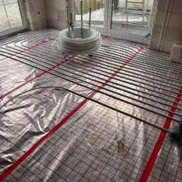 RDK DEVELOER - Instalacja ogrzewania podłogowego w nowo wznoszonym budynku, rury ułożone na folii z czerwoną siatką, białe ściany z pustaków, widok na zewnątrz przez okno.