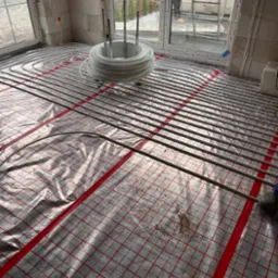 Instalacja ogrzewania podłogowego w nowo wznoszonym budynku, rury ułożone na folii z czerwoną siatką, białe ściany z pustaków, widok na zewnątrz przez okno.