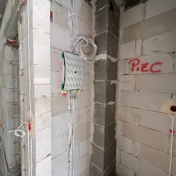 RDK DEVELOER - Wnętrze domu w trakcie budowy: ściany z białych bloczków, piony instalacji elektrycznej z puszkami, narożnik wymurowany z szarych bloczków, napis 'PIEC' na ścianie.