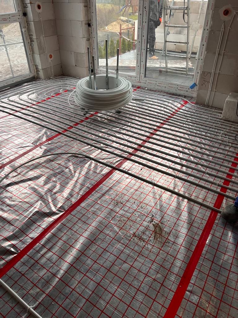 Instalacja ogrzewania podłogowego w nowo wznoszonym budynku, rury ułożone na folii z czerwoną siatką, białe ściany z pustaków, widok na zewnątrz przez okno.