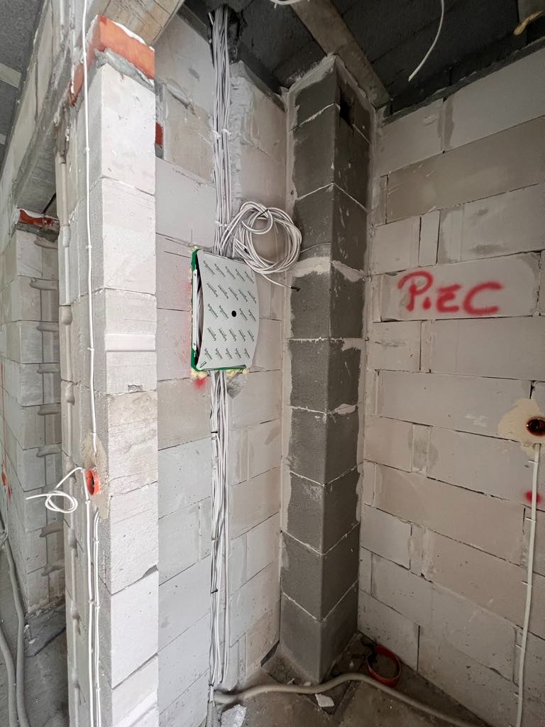 Wnętrze domu w trakcie budowy: ściany z białych bloczków, piony instalacji elektrycznej z puszkami, narożnik wymurowany z szarych bloczków, napis 'PIEC' na ścianie.