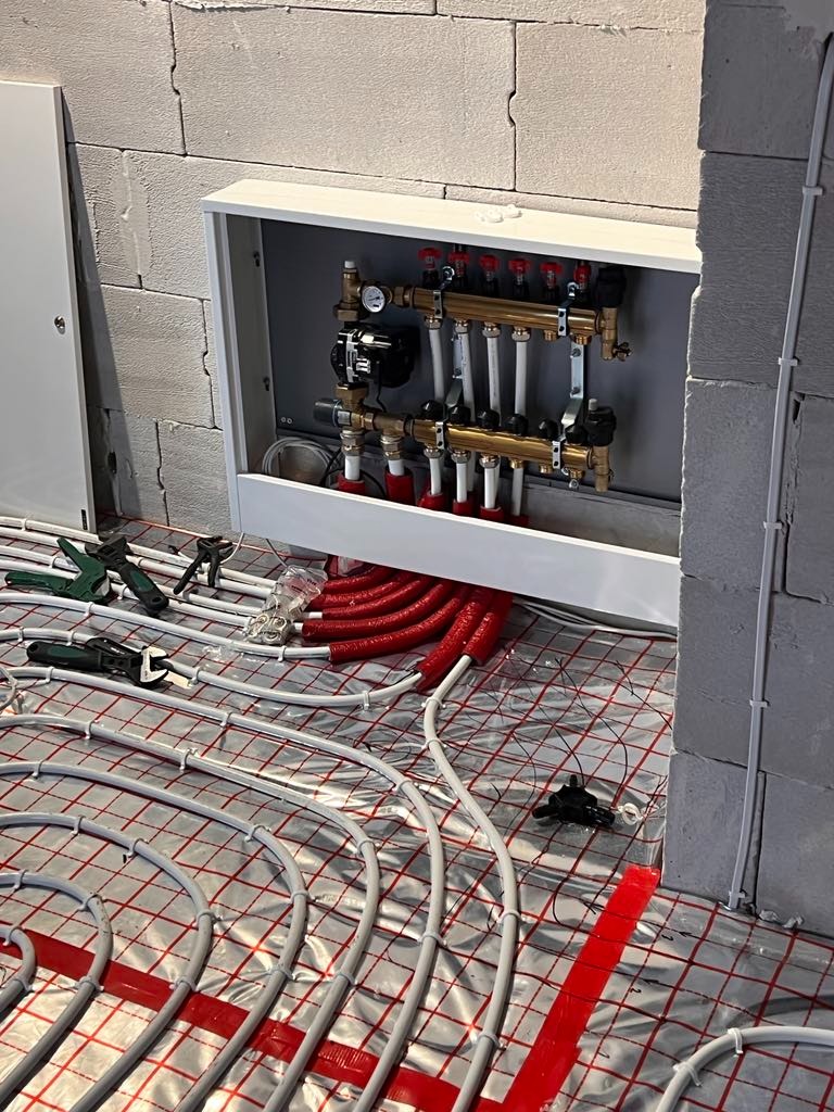 Montaż ogrzewania podłogowego w surowym stanie: rozdzielacz z zaworami, rury grzewcze na folii z siatką, narzędzia montażowe obok.