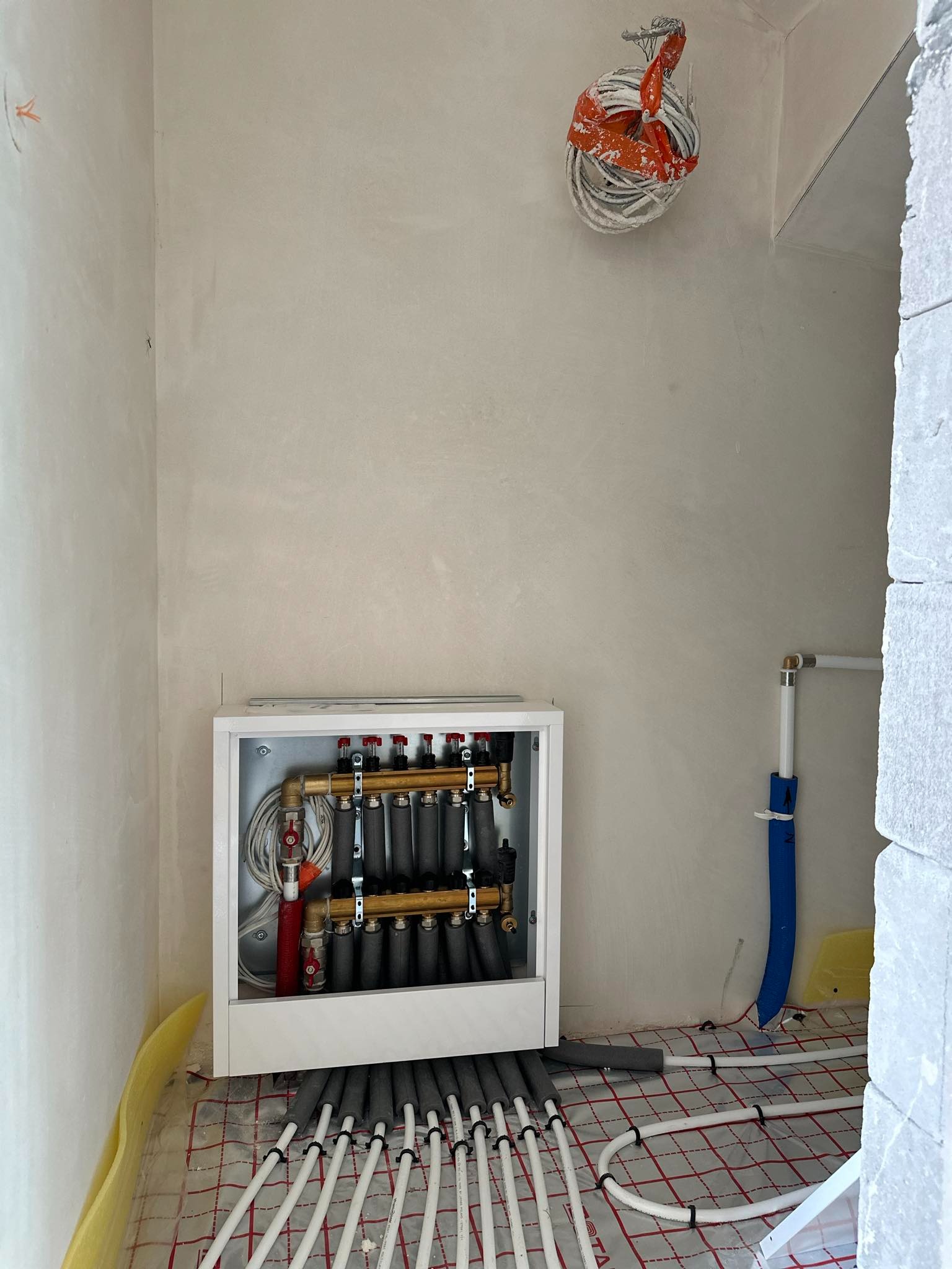 Rozdzielacz ogrzewania podłogowego w szafce, rury rozprowadzone na macie grzewczej w surowym pomieszczeniu z otynkowanymi ścianami i wiązką kabli pod sufitem.