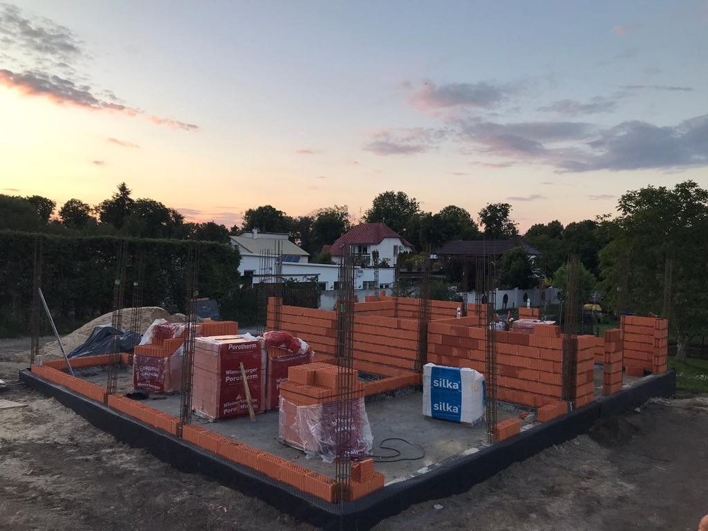 Wznoszenie ścian z czerwonej cegły na fundamencie, widoczne zbrojenia pionowe i paczki cegieł Porotherm i Silka, w tle zabudowa jednorodzinna i zachodzące słońce.