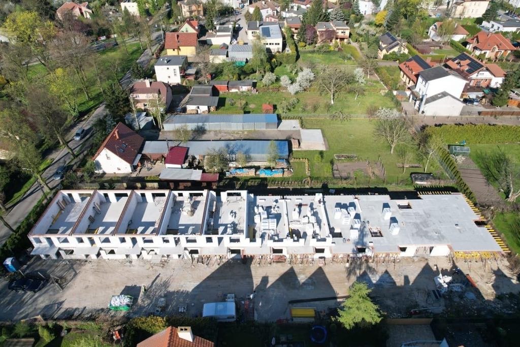 Widok z lotu ptaka na budowę szeregową domów w stanie surowym otwartym, z widocznymi ścianami nośnymi i fundamentami, na tle zieleni i zabudowy mieszkalnej.