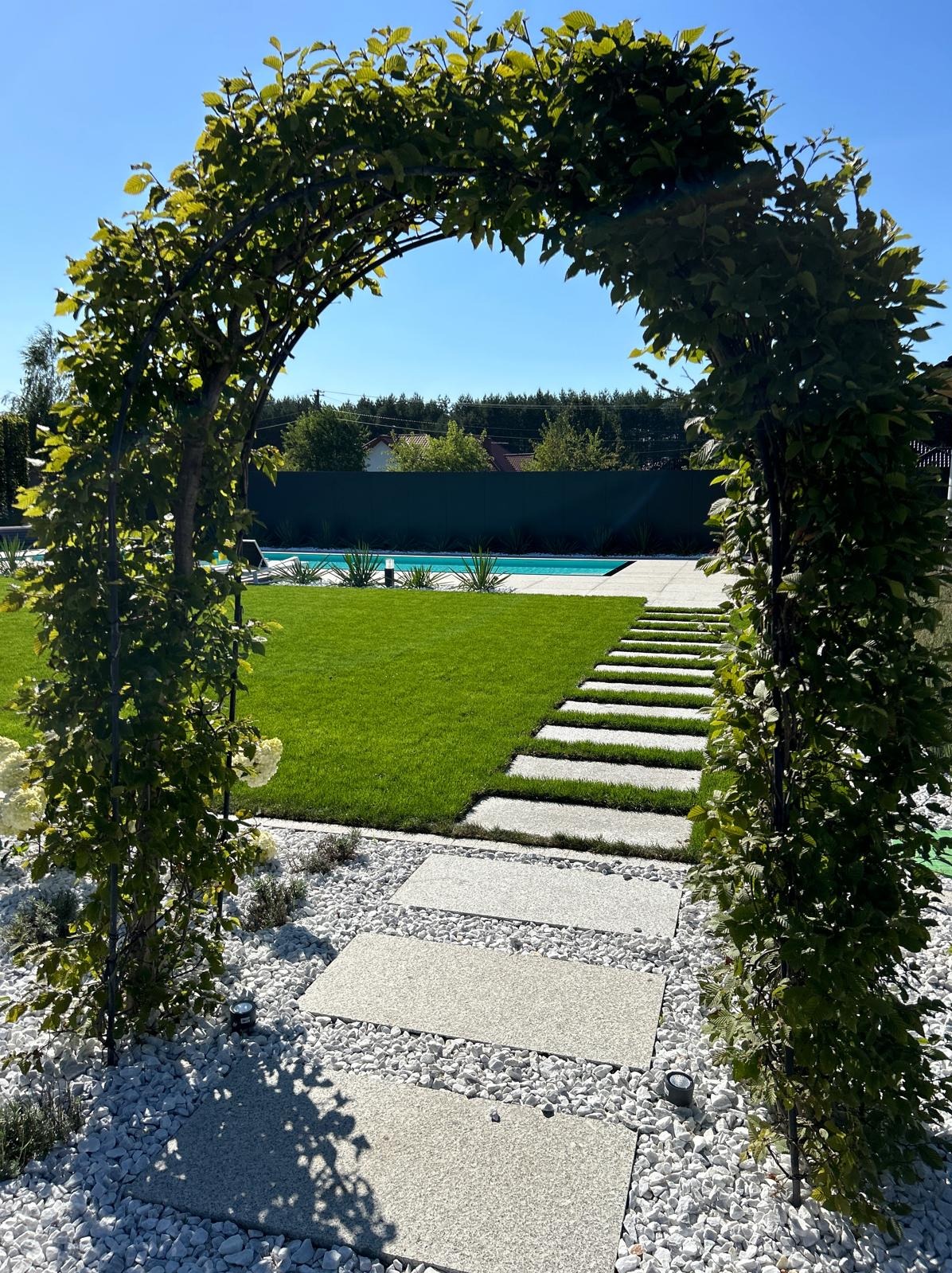 Arka z pnących roślin prowadząca do wypielęgnowanego trawnika z basenem w tle, ścieżka z płyt ułożonych w trawie, dekoracyjny żwir.