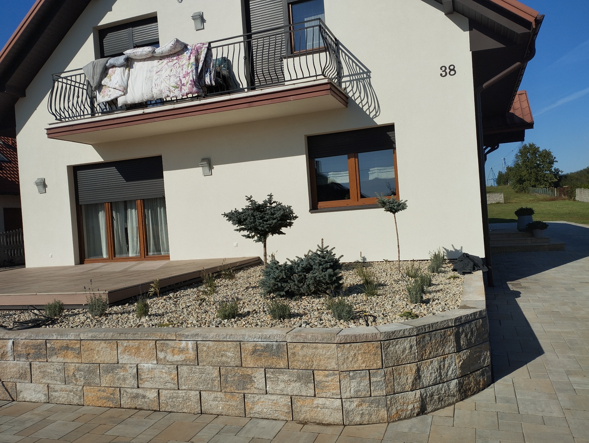 Ogródek skalny z niskimi iglakami i kamieniami, oddzielony od chodnika murkiem z bloków betonowych, w tle dom z balkonem, na którym suszy się pościel.