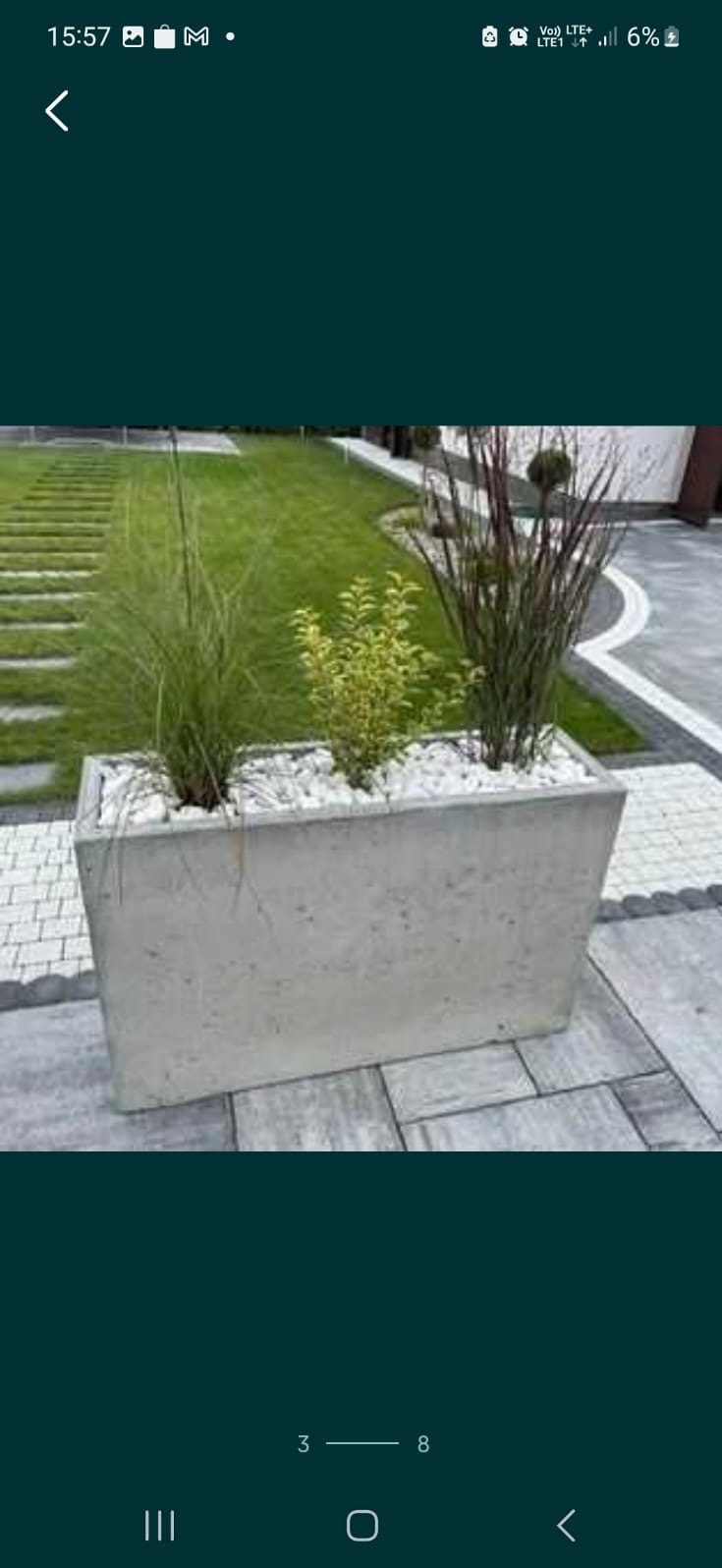 Betonowa donica z ozdobnymi kamieniami i nasadzeniami traw ozdobnych oraz niskiego krzewu na tle zadbanego trawnika z kamiennymi ścieżkami.