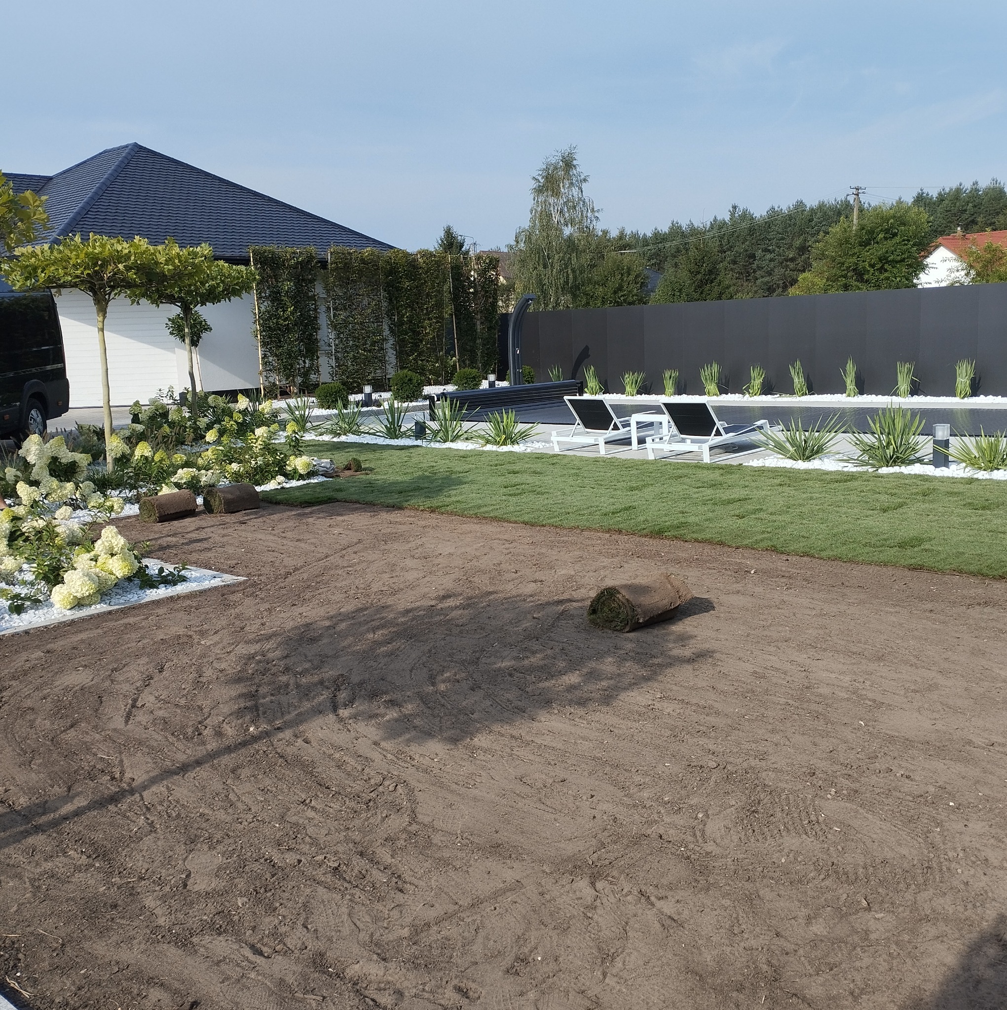 Fragment ogrodu z ułożoną trawą z rolki, widoczne dwie rolki trawy, rabaty z białymi kamieniami i roślinami, w tle czarny płot, leżaki i drzewa.