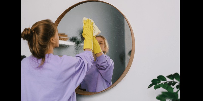 Okrągłe lustro czyszczone ściereczką przez osobę w żółtych rękawicach, widoczne odbicie osoby, roślina w tle.