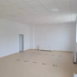 Puste, jasne biuro z białymi ścianami, podwieszanym sufitem i beżową podłogą z kwadratowymi gniazdami podłogowymi, grzejniki przy ścianach.