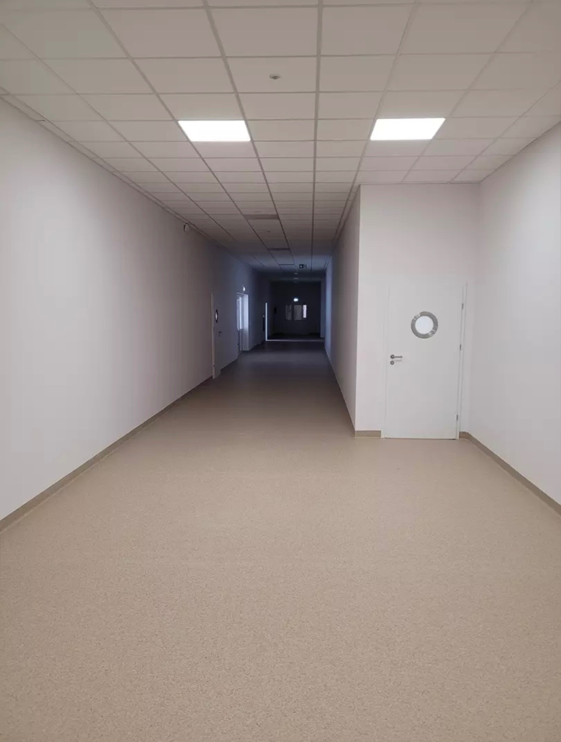 Długi, pusty korytarz z białymi ścianami, podwieszanym sufitem z kwadratowymi lampami i beżową podłogą z wykładziny PCV; drzwi z okrągłym oknem po prawej stronie.