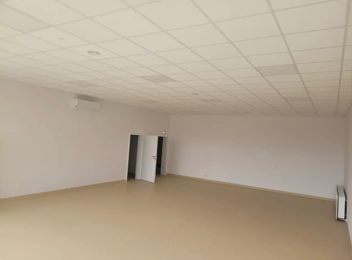 Puste, jasne pomieszczenie biurowe z białymi ścianami, beżową podłogą, podwieszanym sufitem kasetonowym i klimatyzatorem ściennym, przygotowane do aranżacji.