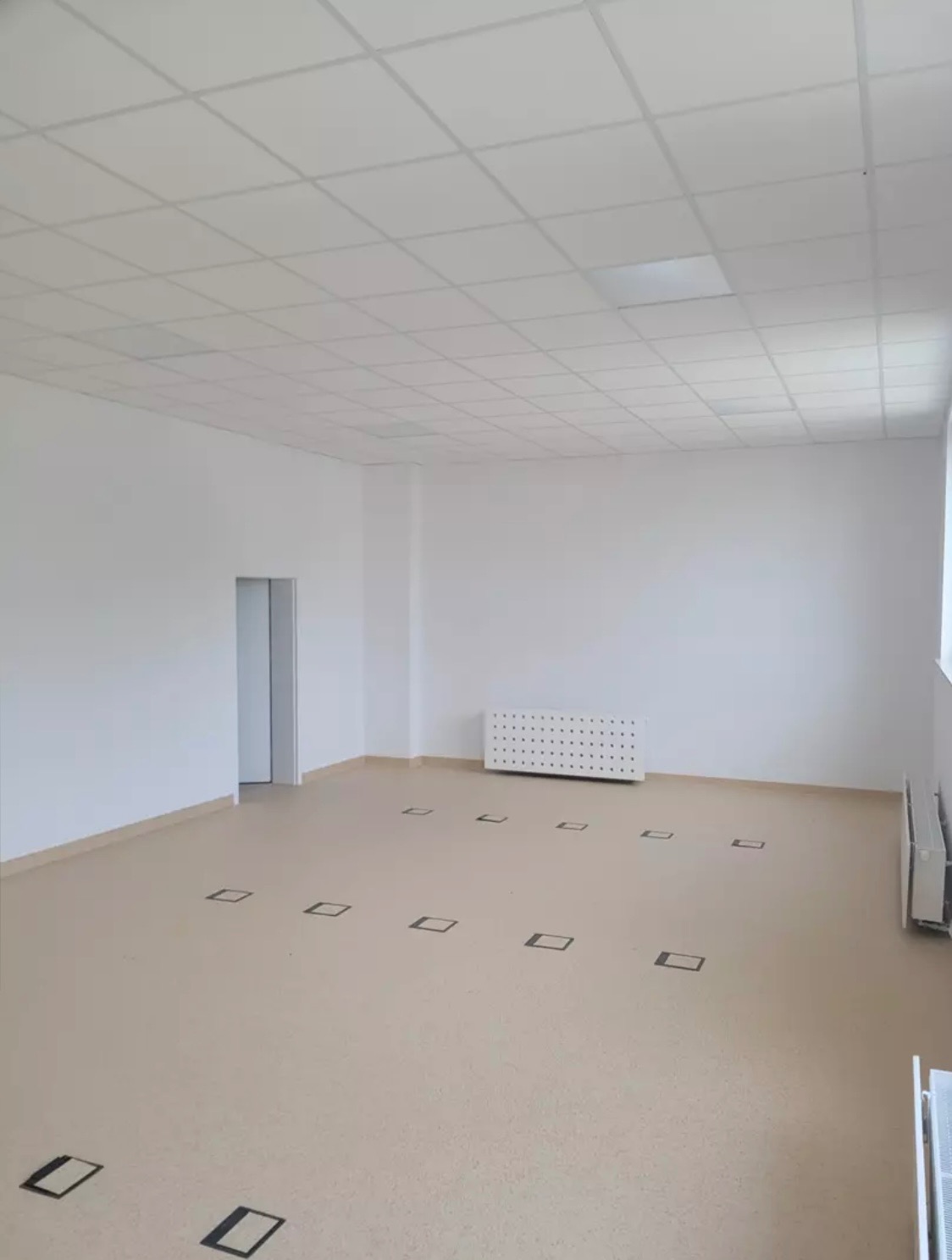 Puste, jasne biuro z białymi ścianami, podwieszanym sufitem i beżową podłogą z kwadratowymi gniazdami podłogowymi, grzejniki przy ścianach.