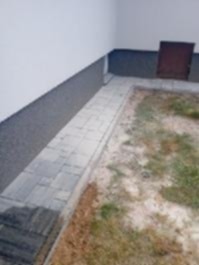 Szara kostka brukowa obramowująca rabatę z suchą trawą przy białej ścianie budynku z ciemnoszarą lamperią. Ujęcie z góry, widoczny fragment brązowych drzwi.