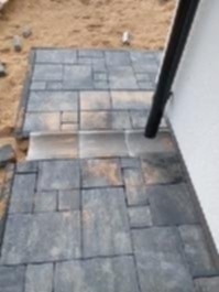 Ułożona kostka brukowa w odcieniach szarości i beżu, z widocznym odpływem liniowym i rurą spustową przy białej ścianie budynku. Piasek w tle.