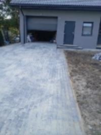 Szara kostka brukowa na podjeździe do garażu z otwartą bramą. Widoczne obrzeża i fragment budynku z szarą elewacją. Ujęcie z niskiej perspektywy.