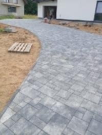 Ścieżka z szarej kostki brukowej prowadząca do domu z białym garażem. Paleta leży na piasku obok ścieżki. Widok z góry, lekko zamazane tło.