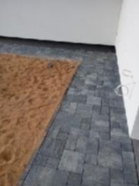 Szara kostka brukowa ułożona w geometryczny wzór, z brązowym piaskiem na części powierzchni. Widok z góry, blisko ściany.