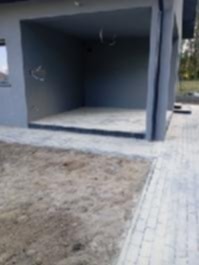 Surowy stan podjazdu z kostki brukowej przed garażem, widoczne przygotowane podłoże i fragment ułożonej nawierzchni. Szary budynek w tle.