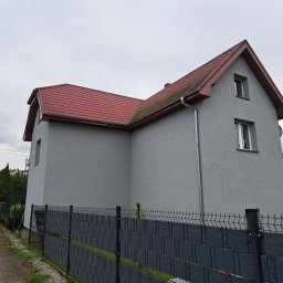 Ernest steczkowski - Dom jednorodzinny po remoncie elewacji, pomalowany na szaro z czerwoną dachówką, widoczny zza czarnego ogrodzenia. Nowe rynny.