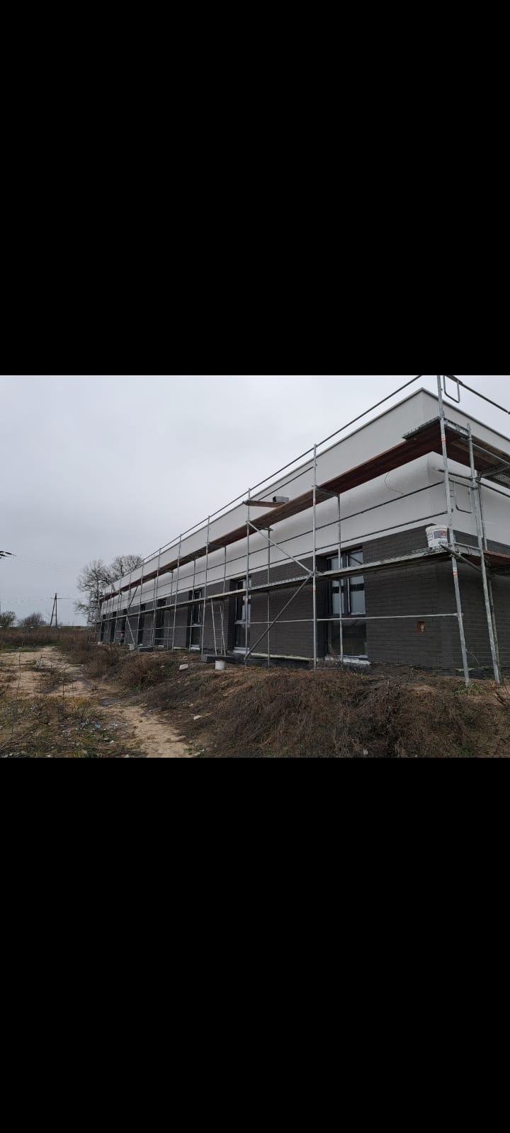 Budynek w trakcie prac elewacyjnych, z widocznymi rusztowaniami i częściowo wykończoną fasadą w odcieniach szarości i bieli. Prace w toku, widoczne materiały budowlane.