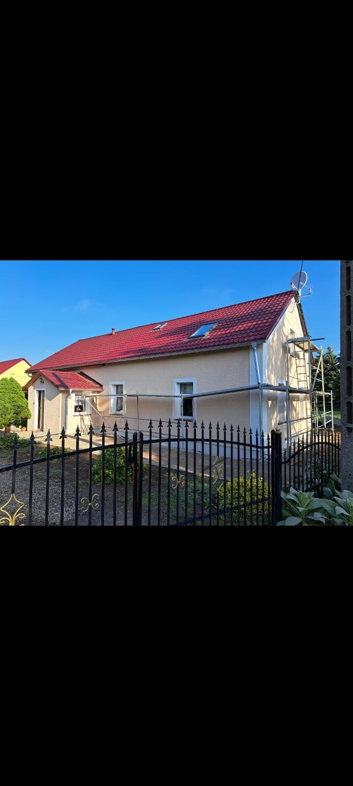 Dom jednorodzinny z czerwoną dachówką, częściowo otoczony rusztowaniem, sugerujący prace elewacyjne w toku. Widoczny fragment ozdobnego, metalowego ogrodzenia.