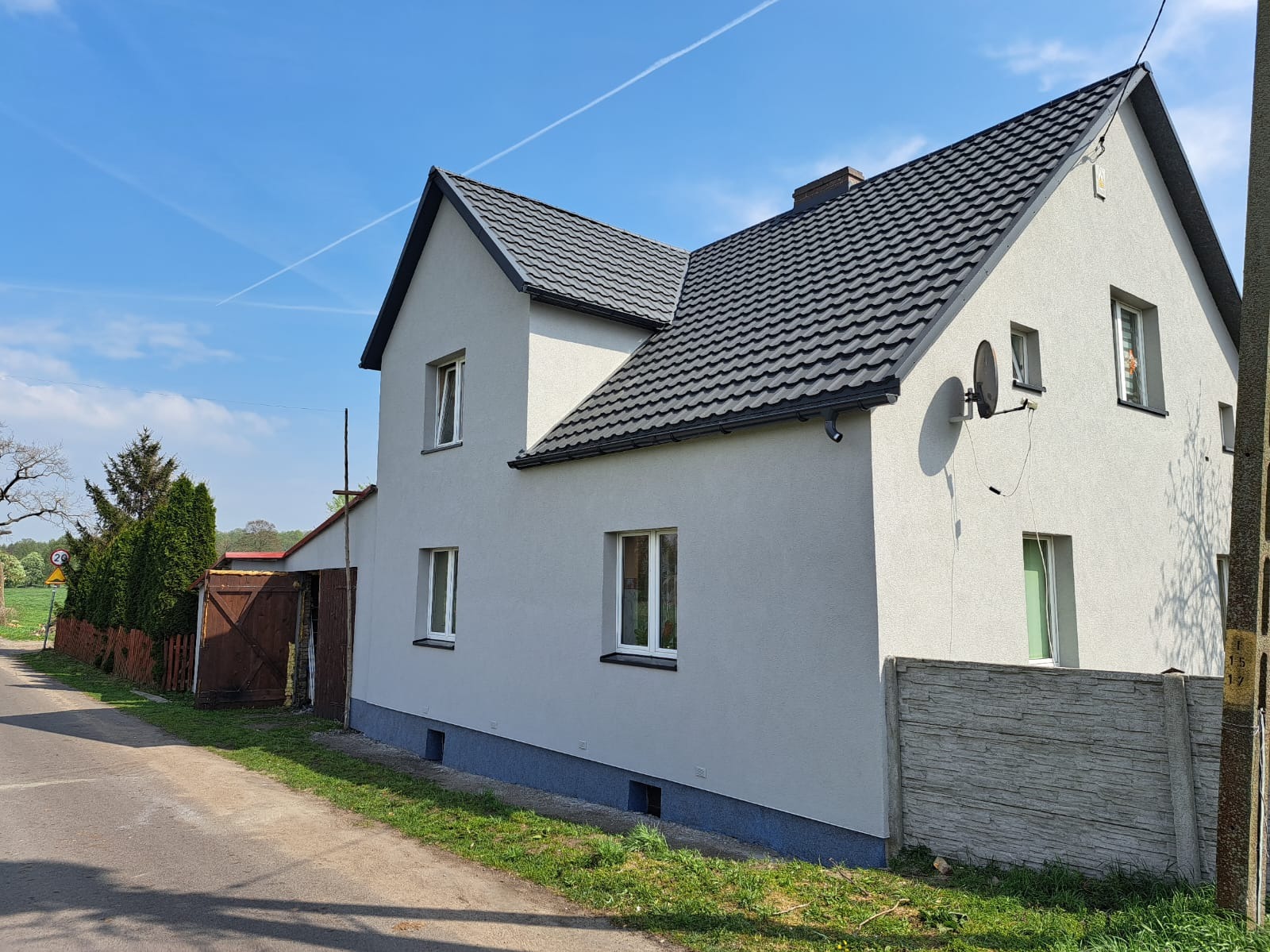 Dwukondygnacyjny dom z szarą elewacją i ciemnym dachem, widziany z boku, z anteną satelitarną na ścianie. Ujęcie w słoneczny dzień, z fragmentem drogi i ogrodzenia.