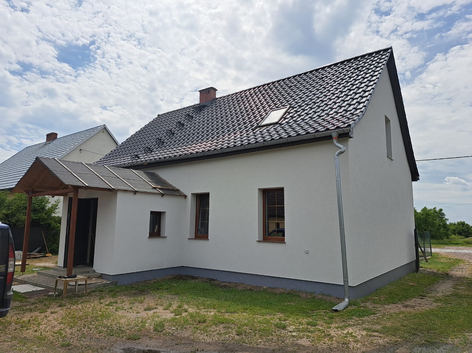 Biały dom jednorodzinny z ciemną dachówką i drewnianą wiatą przy wejściu, po remoncie elewacji. Widoczne okna z brązowymi ramami i szara podmurówka.