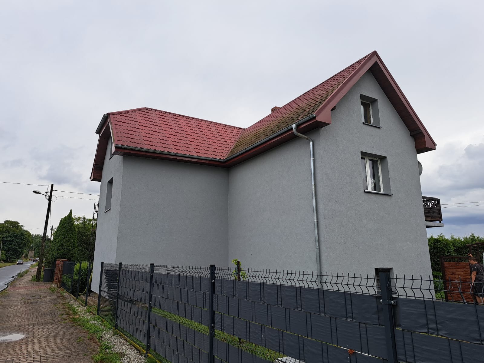 Dom jednorodzinny po remoncie elewacji, pomalowany na szaro z czerwoną dachówką, widoczny zza czarnego ogrodzenia. Nowe rynny.