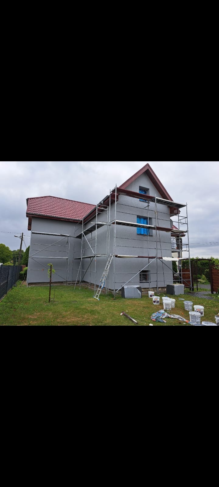 Dom jednorodzinny w trakcie malowania elewacji na szaro, z rozstawionymi rusztowaniami i widocznymi pojemnikami z farbą na trawniku. Czerwony dach.