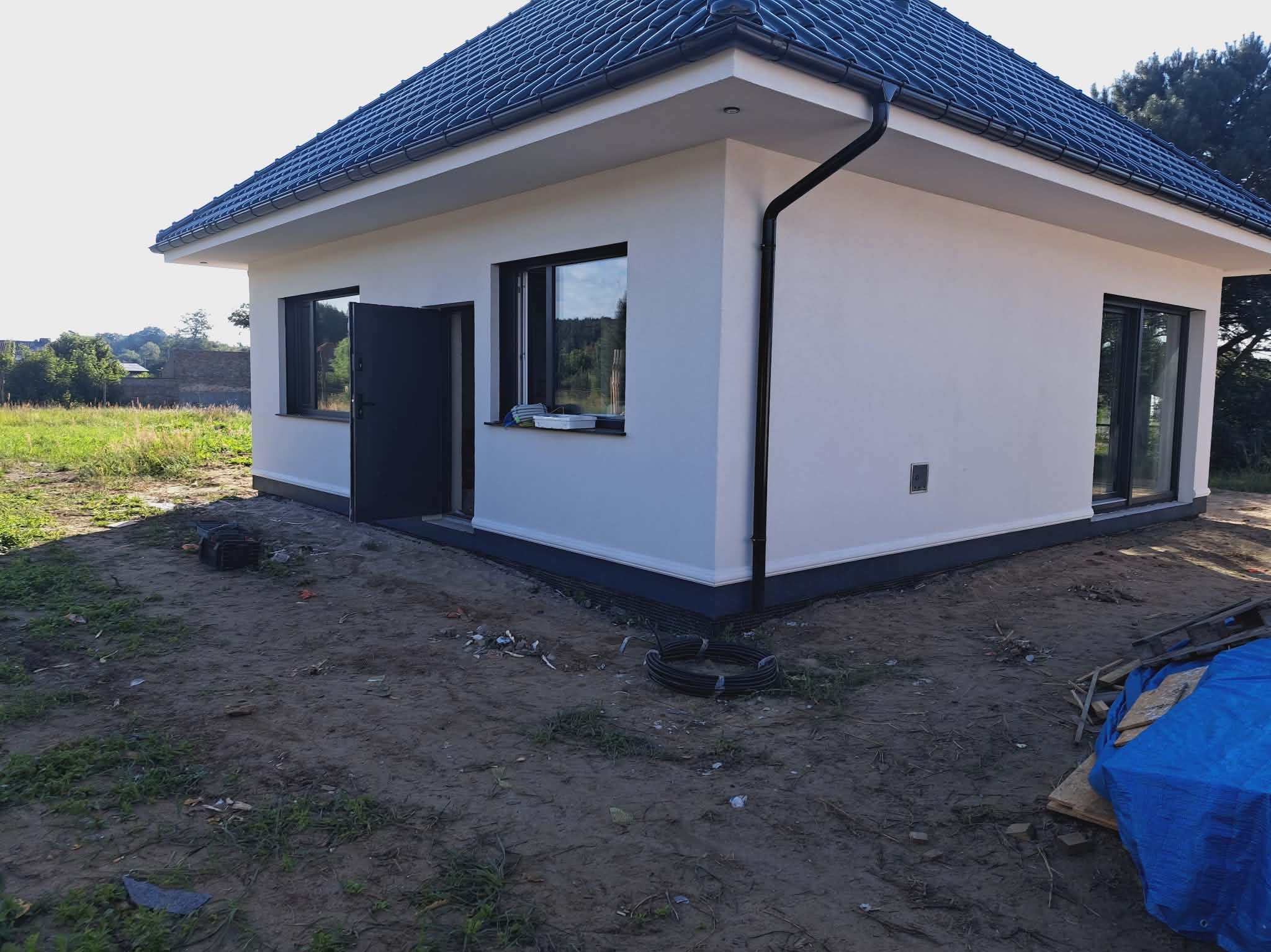 Biały, parterowy dom z ciemną dachówką i czarnymi oknami. Widoczne wykończone elewacje, rynny i cokół. Otwarta brama wejściowa. Wokół domu ziemia i resztki materiałów budowlanych.