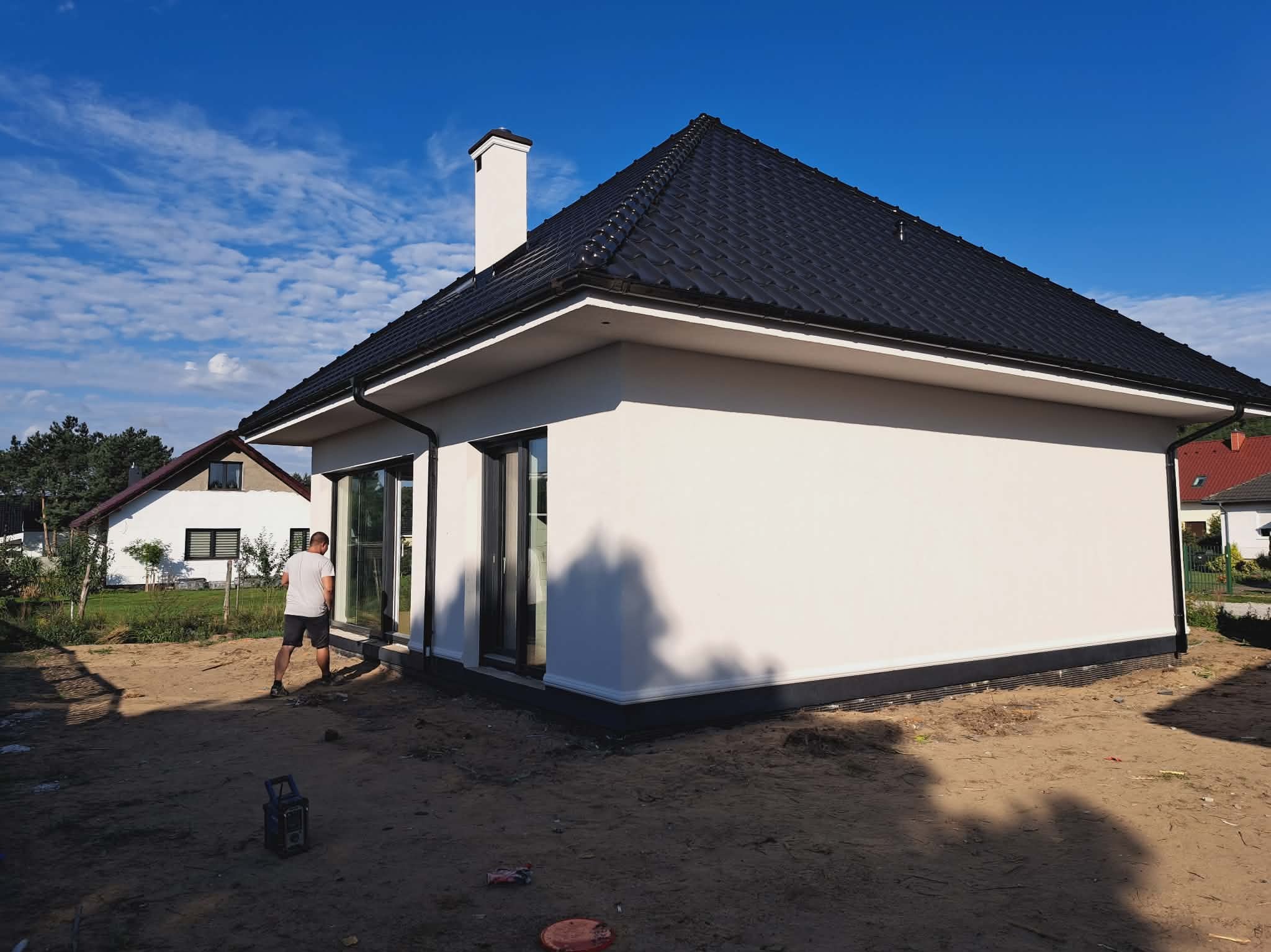 Nowoczesny dom jednorodzinny z białą elewacją i czarnym dachem, widoczny z boku. Na pierwszym planie piasek, w tle fragment sylwetki mężczyzny i sąsiedni budynek.