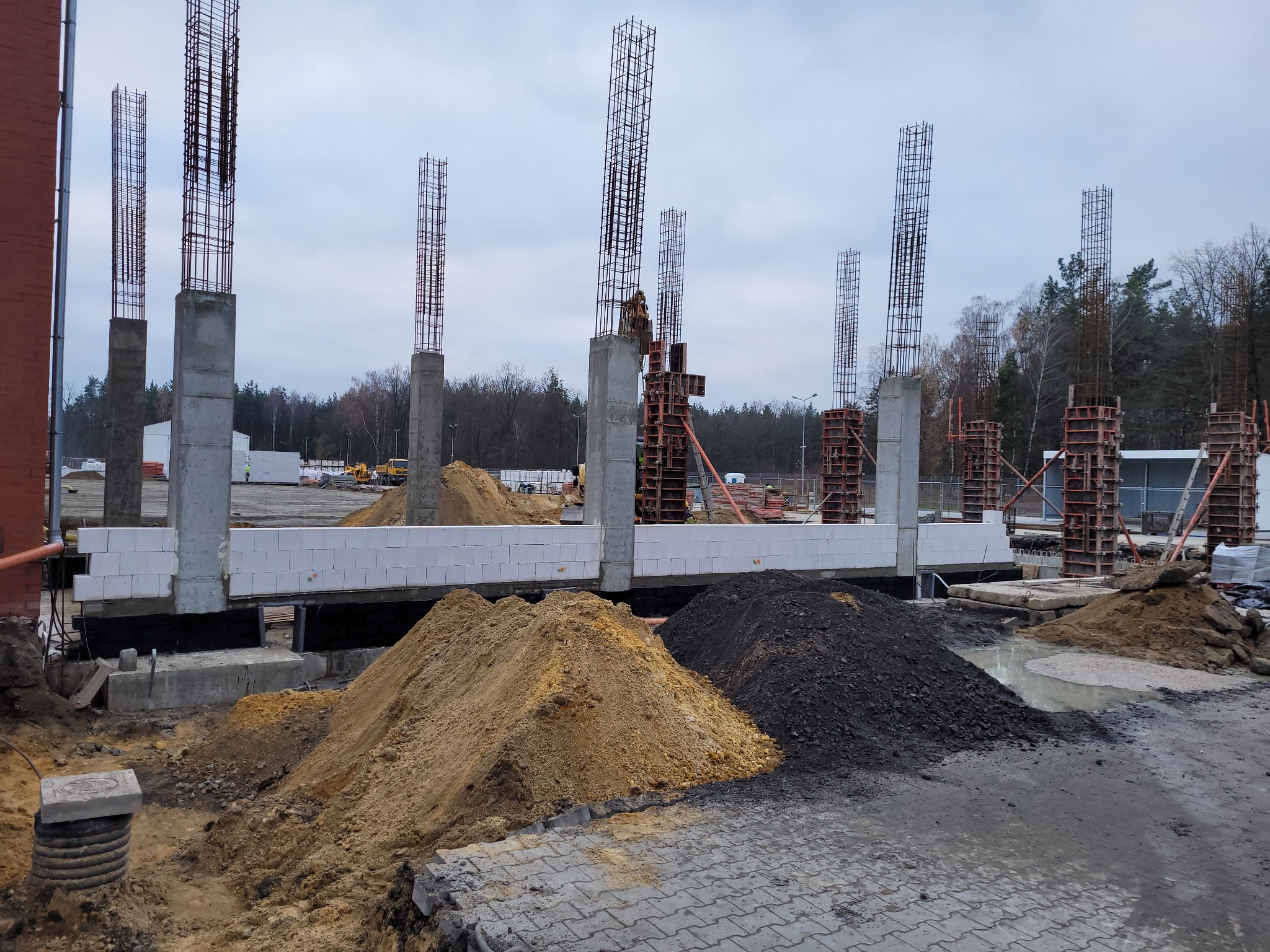 Wczesny etap budowy budynku z widocznymi zbrojeniami kolumn, szalunkami, fundamentami z bloczków betonowych oraz stertami piasku i ziemi na placu budowy w pochmurny dzień.