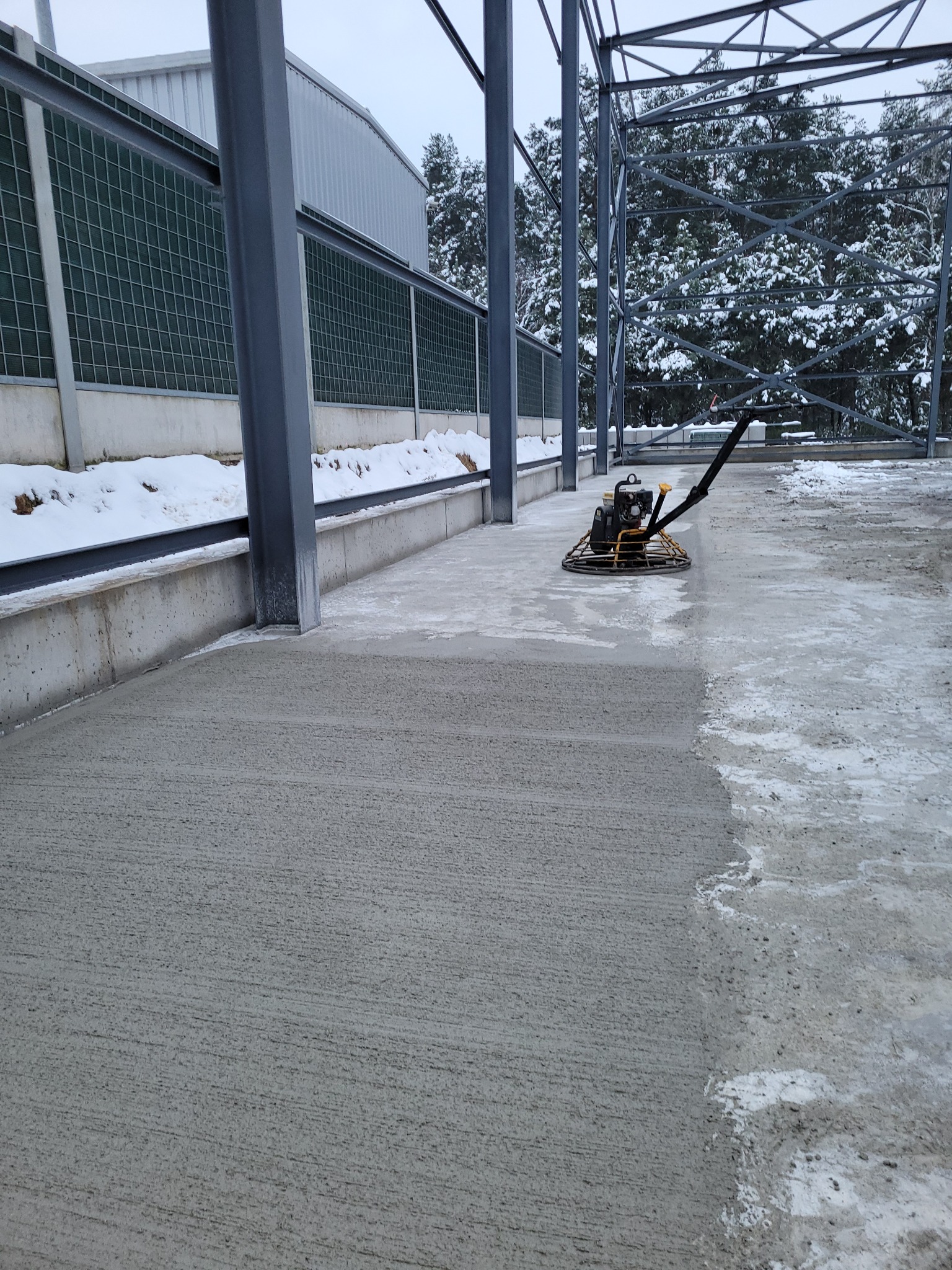 Zacieraczka mechaniczna do betonu w trakcie pracy na szarej, betonowej posadzce wewnątrz konstrukcji stalowej, z widocznym śniegiem na zewnątrz.