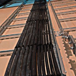 Usługi elektryczne Kuczyński Sebastian - Równoległe wiązki przewodów elektrycznych, czarne kable ułożone w korytku kablowym, widoczne uchwyty mocujące i metalowa konstrukcja wsporcza, instalacja w budynku w trakcie realizacji.