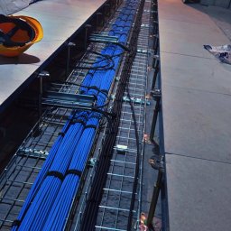 Usługi elektryczne Kuczyński Sebastian - Długi, wykopany kanał z metalową siatką, wypełniony równolegle ułożonymi niebieskimi i czarnymi kablami elektrycznymi, widok z góry, z kaskiem ochronnym w rogu kadru.