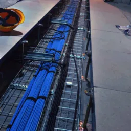 Długi, wykopany kanał z metalową siatką, wypełniony równolegle ułożonymi niebieskimi i czarnymi kablami elektrycznymi, widok z góry, z kaskiem ochronnym w rogu kadru.