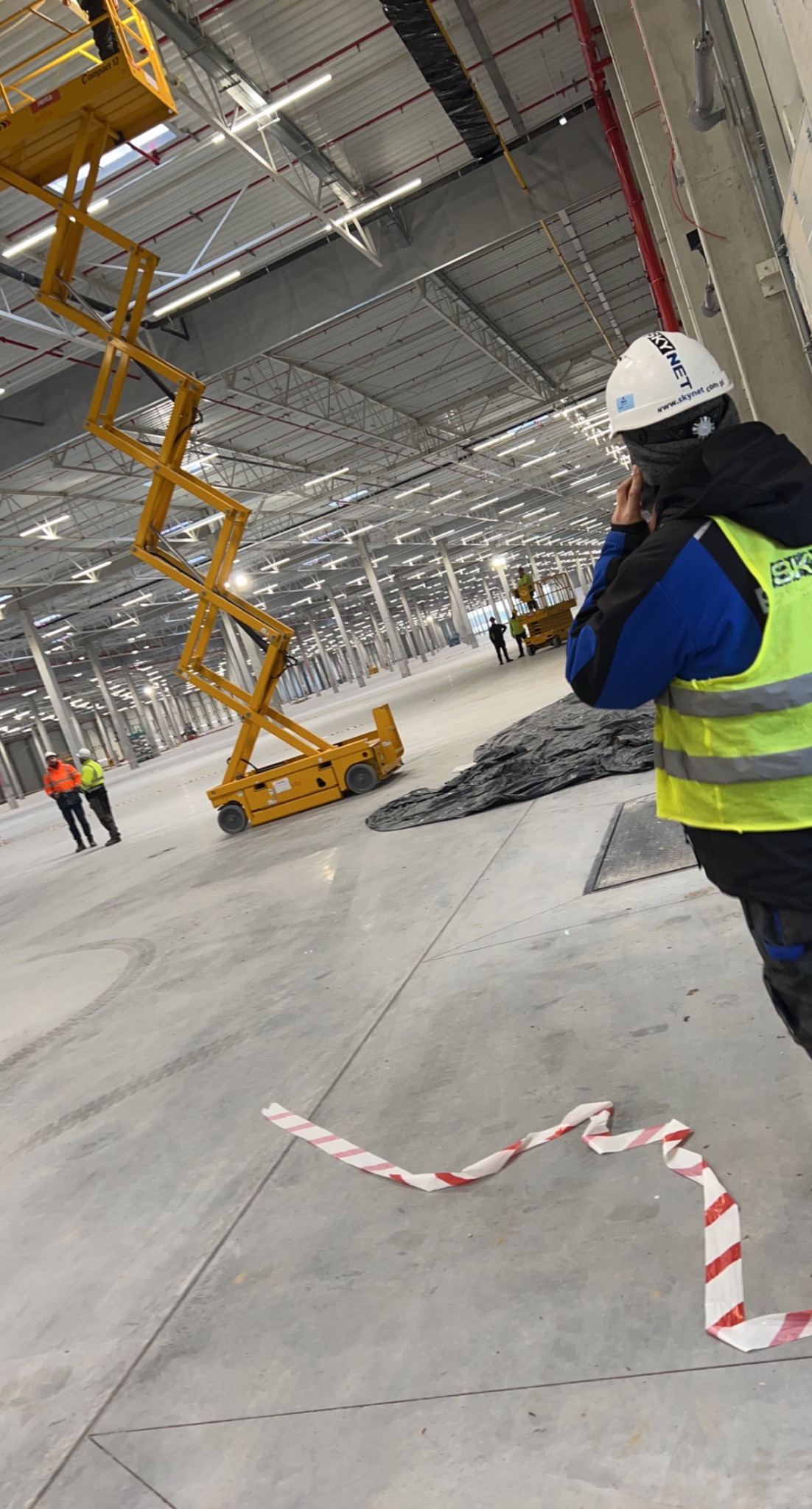 Pracownik w kasku i kamizelce odblaskowej rozmawia przez telefon na tle hali produkcyjnej z podnośnikiem nożycowym i rozciągniętą taśmą ostrzegawczą na podłodze.