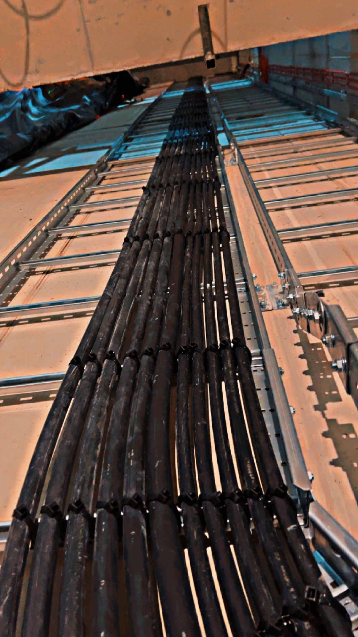 Równoległe wiązki przewodów elektrycznych, czarne kable ułożone w korytku kablowym, widoczne uchwyty mocujące i metalowa konstrukcja wsporcza, instalacja w budynku w trakcie realizacji.