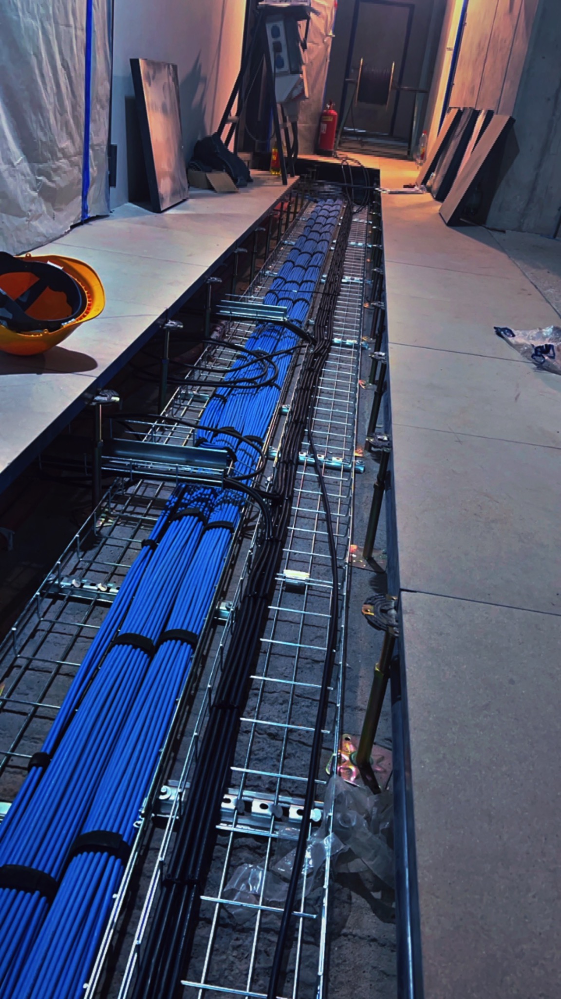 Długi, wykopany kanał z metalową siatką, wypełniony równolegle ułożonymi niebieskimi i czarnymi kablami elektrycznymi, widok z góry, z kaskiem ochronnym w rogu kadru.