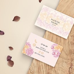 Concept and creation - Dwie wizytówki kwiaciarni z motywem kwiatowym, ułożone na drewnianej desce i beżowym tle z rozrzuconymi płatkami kwiatów. Wizytówki zawierają dane kontaktowe i logo.