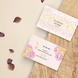 Dwie wizytówki kwiaciarni z motywem kwiatowym, ułożone na drewnianej desce i beżowym tle z rozrzuconymi płatkami kwiatów. Wizytówki zawierają dane kontaktowe i logo.