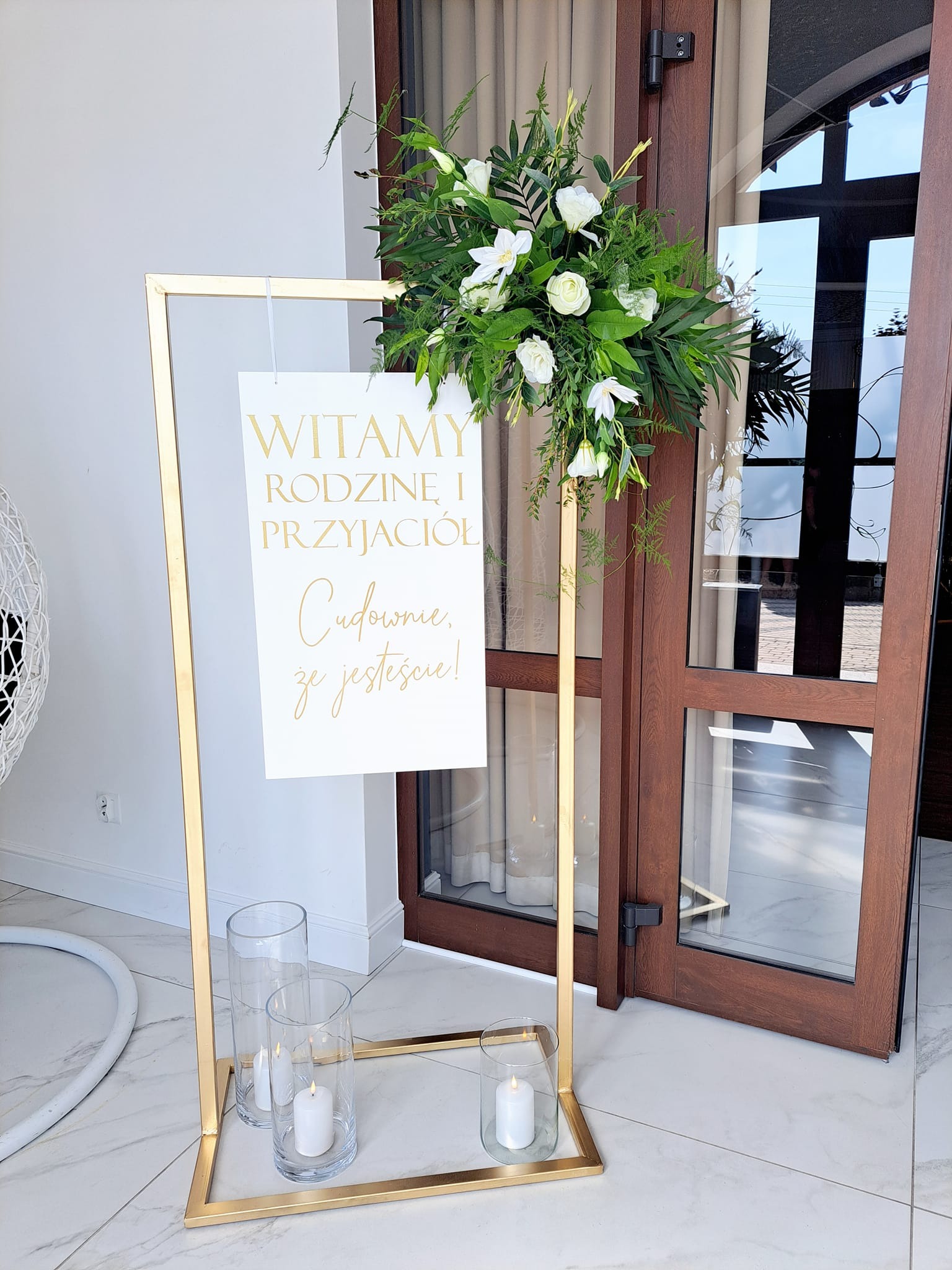 Dekoracja powitalna na ślub: złota rama z napisem 'Witamy Rodzinę i Przyjaciół', kwiatowa kompozycja i świece w szklanych wazonach przy wejściu.