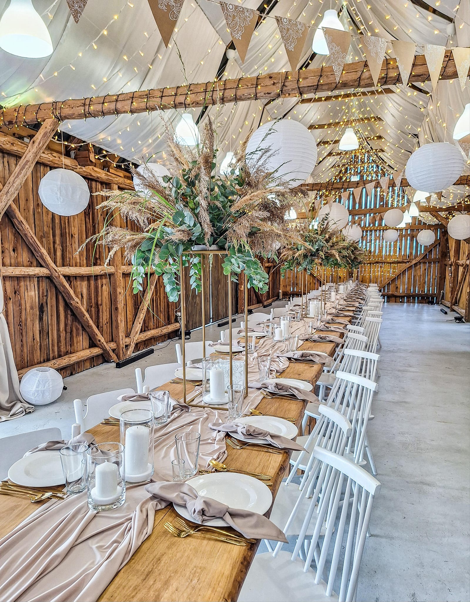 Długi stół weselny z drewnianym blatem, nakryty jasnoróżowym bieżnikiem, białymi talerzami, złotymi sztućcami i beżowymi serwetkami, udekorowany wysokimi kompozycjami z traw pampasowych...