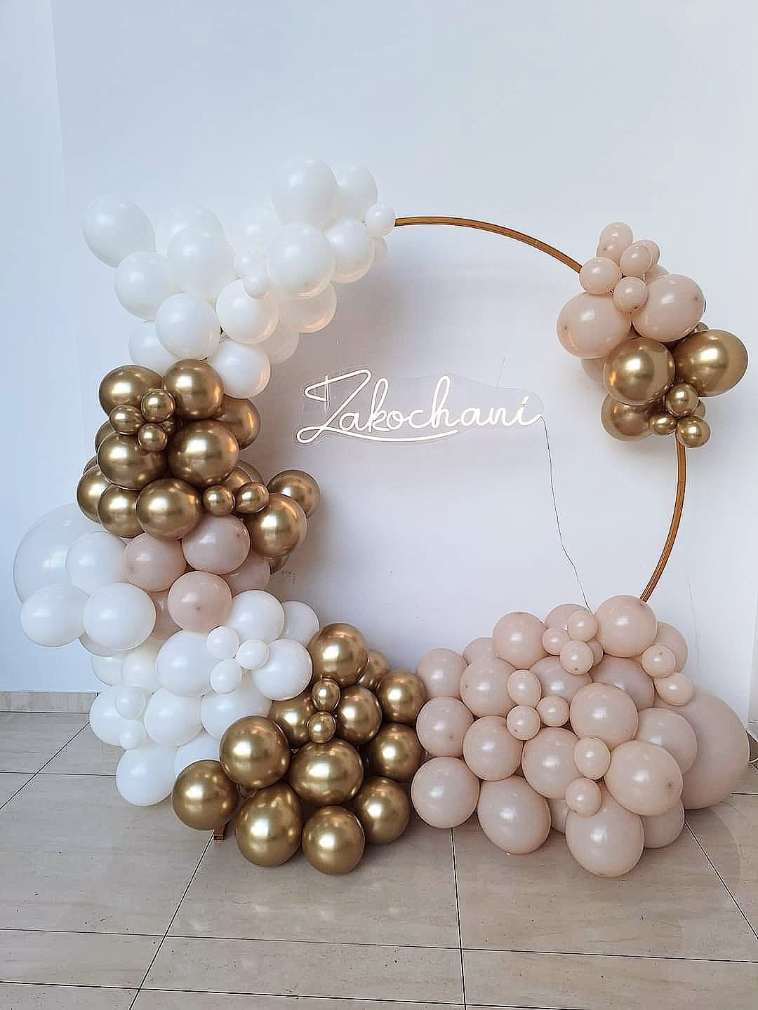 Okrągła, złota rama udekorowana asymetrycznie białymi, złotymi i pastelowo-różowymi balonami, z neonowym napisem 'Zakochani' wewnątrz